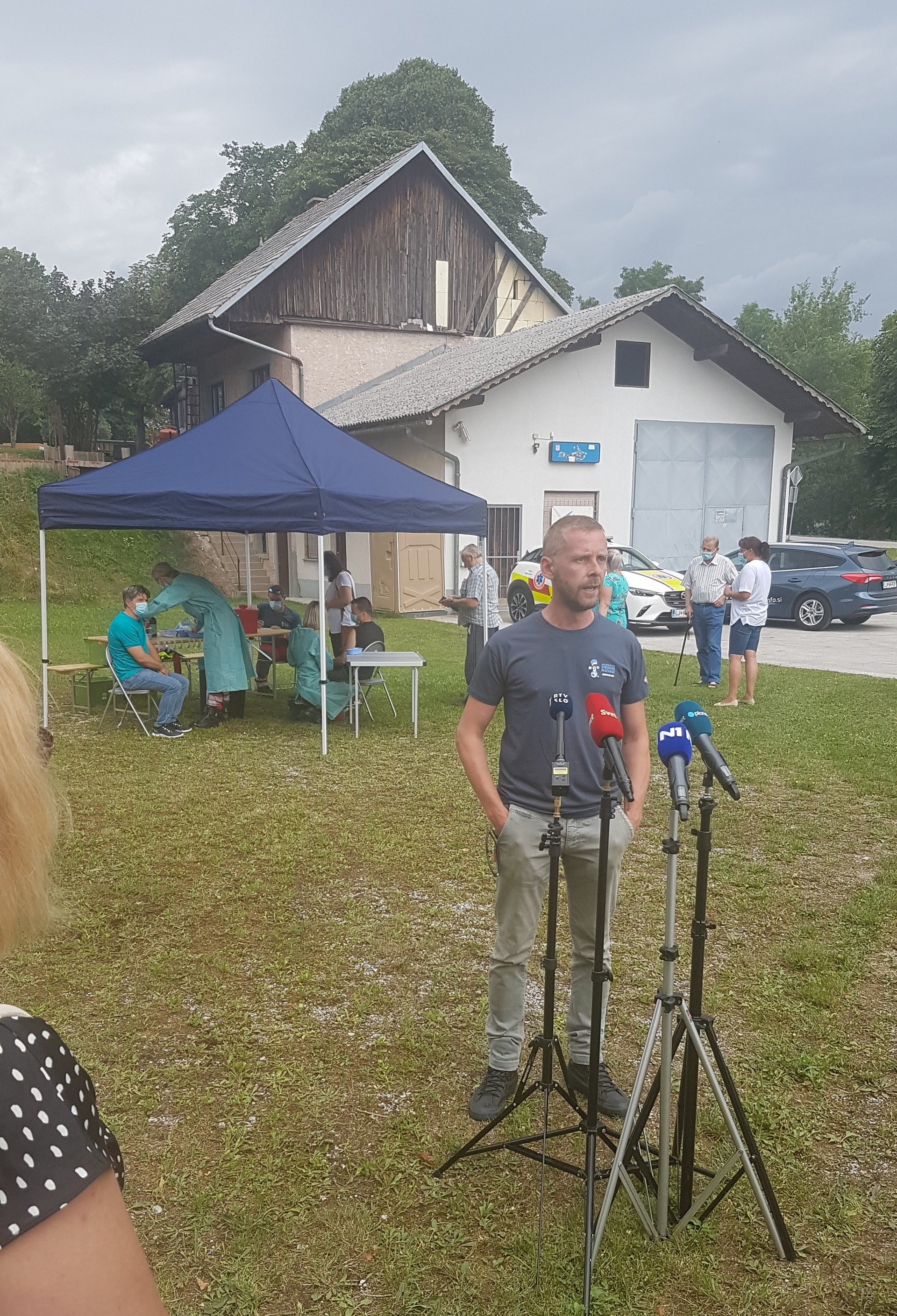 Moški daje izjavo za medije na travniku, ped njim so postavljeni televizijski mikrofoni, za njim je vidna mobilna cepilna enota 