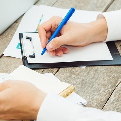 Simbolna slika mize in rok, ki držijo kalkulator v levi in nalivno pero, s katerim piše na papir. Vidijo se še prenosnik, miška in tablica.